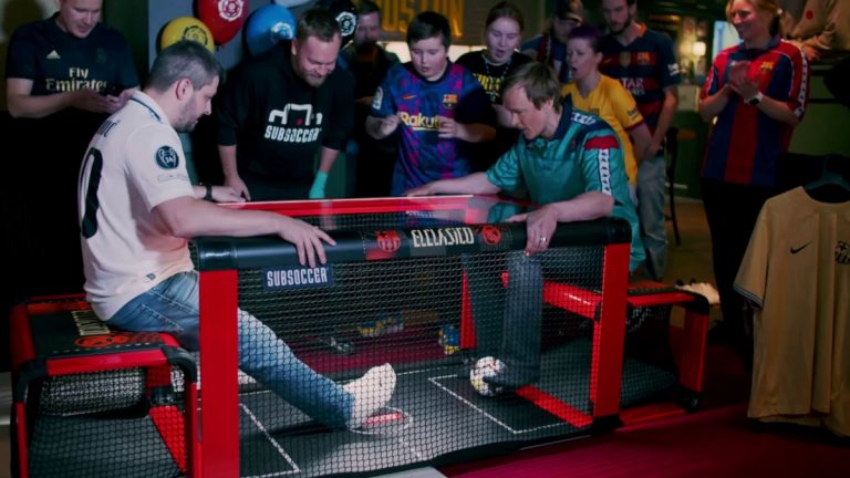 Home 7 El Clasico competition as 2 males play Subsoccer® table football game with crowd watching at event
